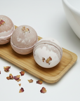 Three bath bombs on a wooden board with dried rose petals, next to a white bowl.