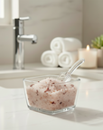 Glass container of soft girl bath salts on a bathroom counter with towels and a candle in the background.