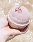 Pink bath bomb held over a pink bath with bubbles